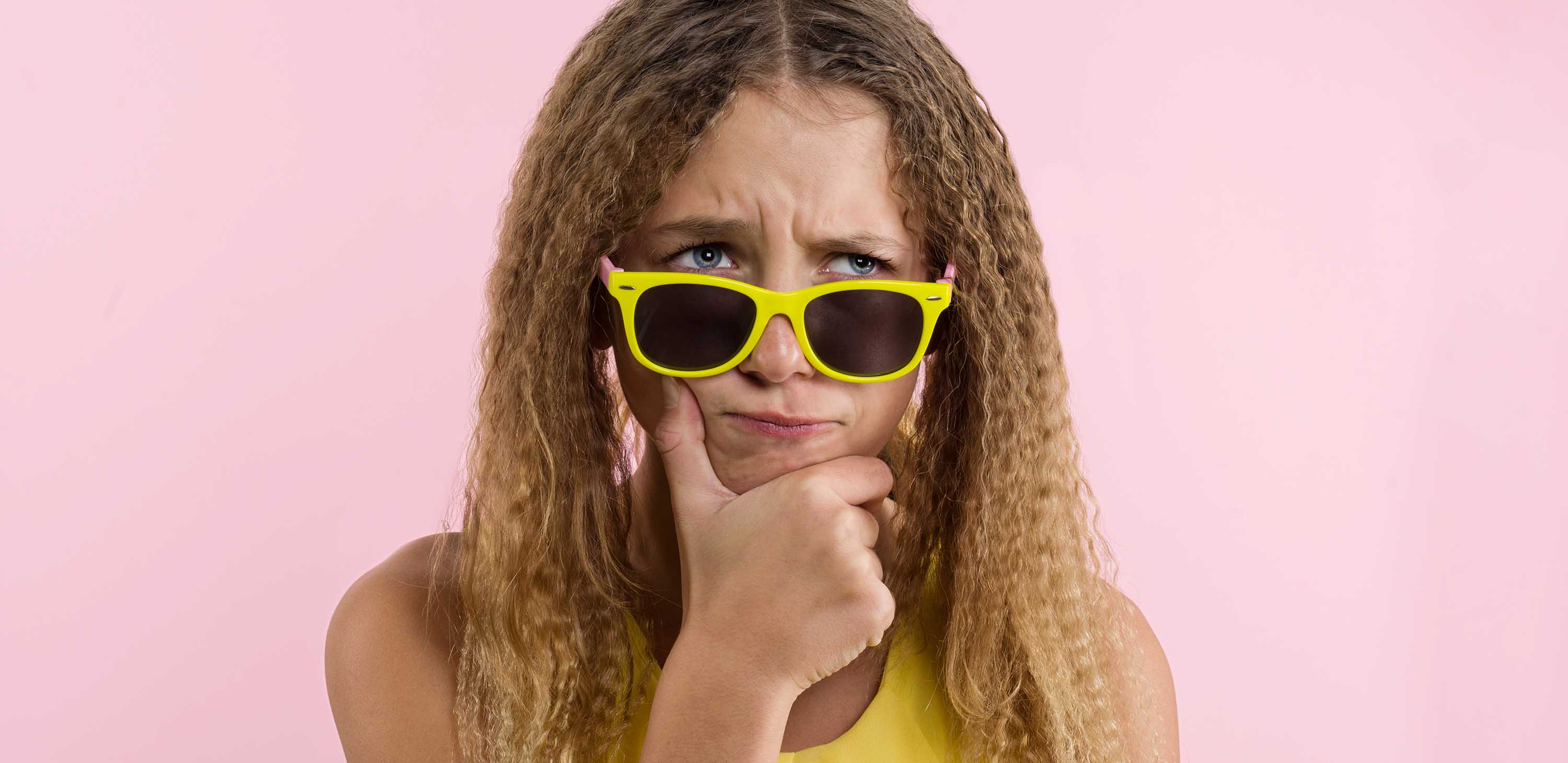 Teenage girl blonde with curly hair frowning, frowning her eyebrow while looking away with placid and thoughtful look. Pensive girl with a puzzled expression on a pink studio background