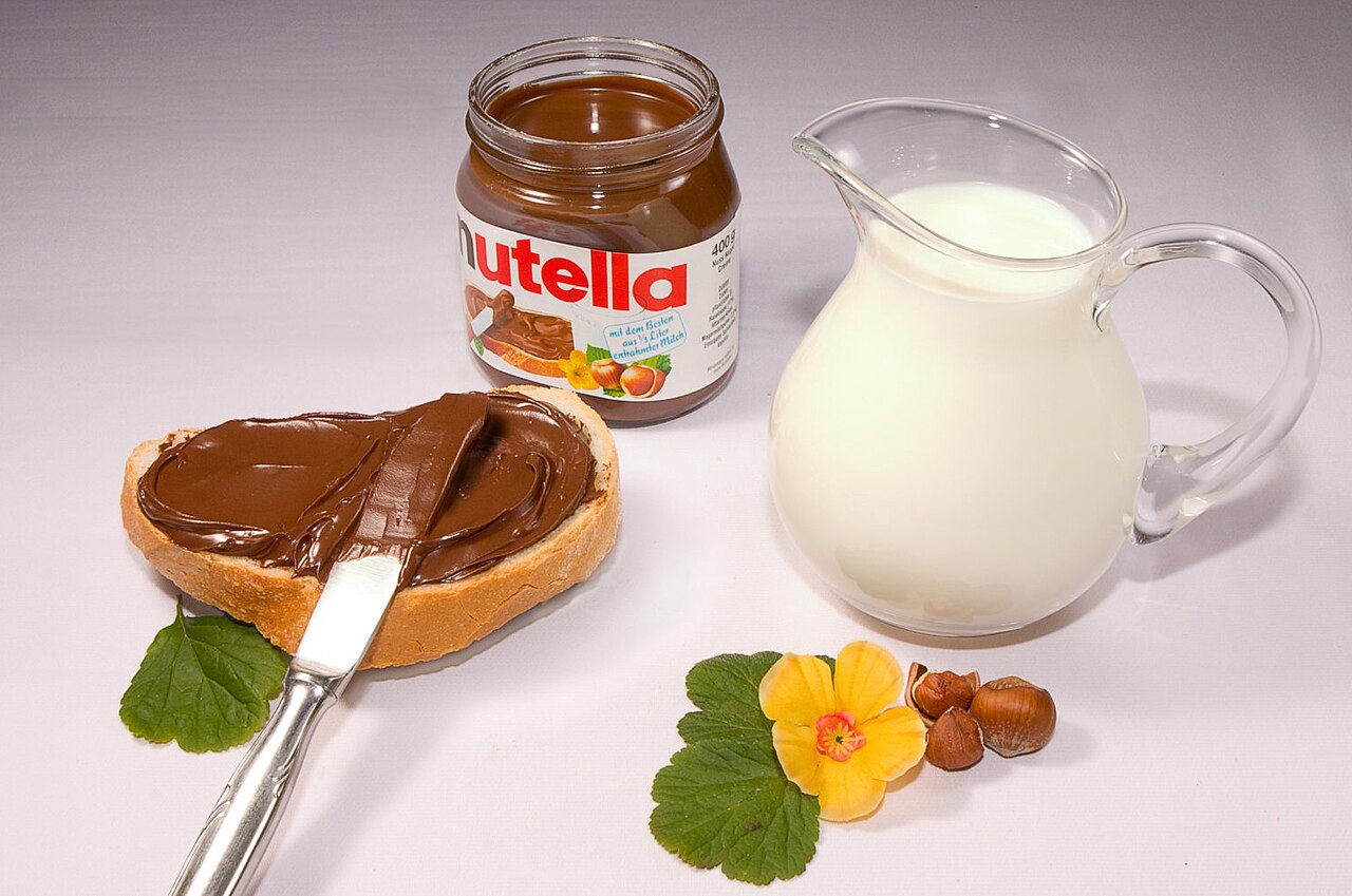An open jar of Nutella beside bread spread with Nutella, a jug of milk and a flower